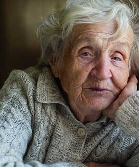 Elderly woman with gray hair resting her head on her hand while sitting indoors in a sweater. - Olive Oil Times
