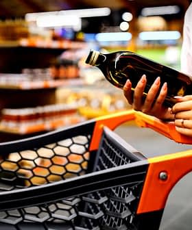 Individual holding a bottle of olive oil while standing next to a shopping cart in a store. - Olive Oil Times