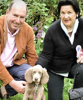 A man and a woman kneeling beside a light brown puppy in a garden setting. - Olive Oil Times