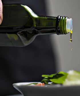 A person pouring olive oil from a dark green bottle onto a fresh salad in a white bowl. - Olive Oil Times