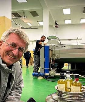 A man smiling in an olive oil processing facility with machinery and bottles of olive oil in the foreground. - Olive Oil Times