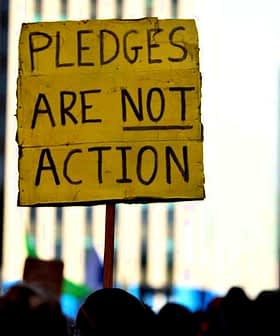 Yellow sign at a protest reading 'Pledges Are Not Action' held by an individual in a crowd. - Olive Oil Times