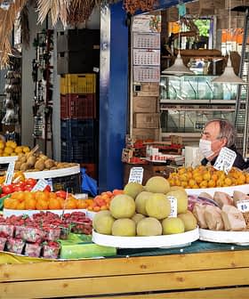Market stall displaying a variety of fruits and vegetables, with a vendor and a customer wearing masks. - Olive Oil Times