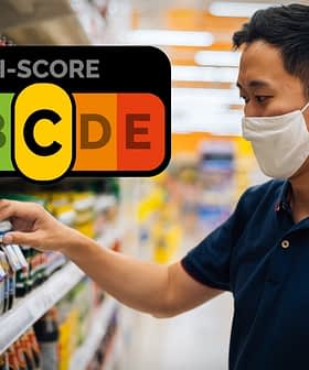 Man wearing a mask examining beverage labels on a supermarket shelf with a Nutri-Score graphic in the background. - Olive Oil Times