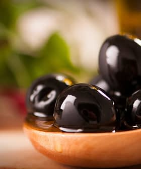 A wooden spoon holding several black olives in olive oil, with a blurred background. - Olive Oil Times