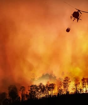 Helicopter dropping water on a large wildfire with flames and smoke in the background. - Olive Oil Times