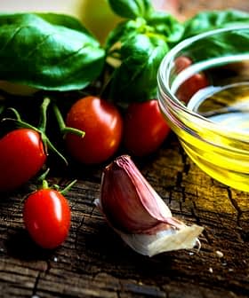 Cherry tomatoes, garlic, fresh basil, and a small bowl of olive oil on a wooden surface. - Olive Oil Times