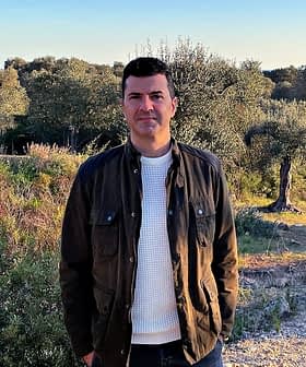 A man wearing a dark jacket and white sweater standing in an olive grove with trees in the background. - Olive Oil Times