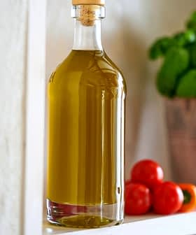 Glass bottle filled with olive oil placed on a shelf next to cherry tomatoes and a basil plant. - Olive Oil Times