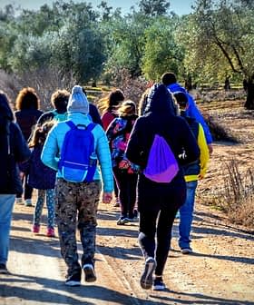 A group of people walking along a dirt path in an olive grove, wearing winter clothing and backpacks. - Olive Oil Times