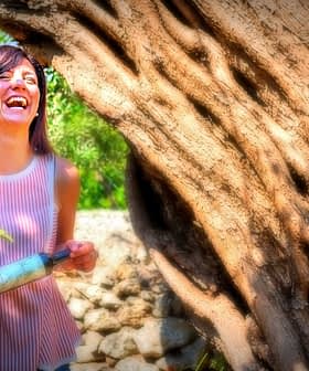 Woman with dark hair laughing while holding a bottle near an olive tree. - Olive Oil Times
