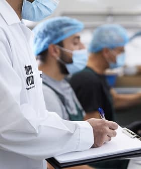 Three workers in a food processing facility wearing masks and hairnets, with one taking notes on a clipboard. - Olive Oil Times