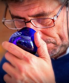 Man with glasses holding a blue glass cup to his nose, appearing to smell it. - Olive Oil Times