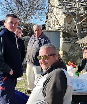 A group of six men gathered around a table outdoors, with stone buildings in the background. - Olive Oil Times
