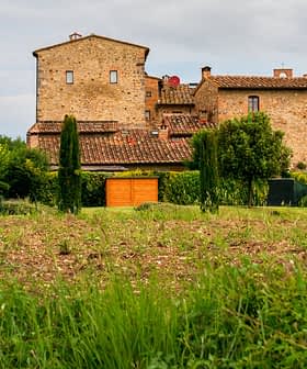 A stone building with a red roof surrounded by greenery and a field in a rural area. - Olive Oil Times