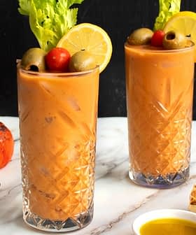 Two tall glasses of tomato-based beverage garnished with celery, olives, and lemon slices on a marble surface. - Olive Oil Times