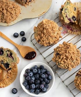 Freshly baked blueberry muffins with a crumb topping on a cooling rack and a plate of blueberries. - Olive Oil Times