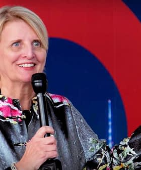 A woman with short blonde hair speaking into a microphone at an event, wearing a floral-patterned jacket. - Olive Oil Times