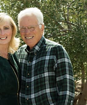 A couple smiling while posing outdoors in front of olive trees, wearing casual clothing. - Olive Oil Times