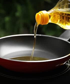A bottle of cooking oil being poured into a red frying pan on a stovetop. - Olive Oil Times