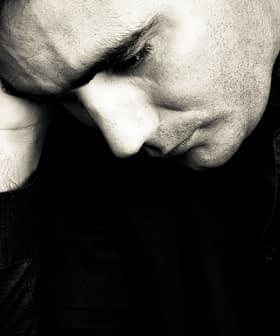 A man in a black shirt resting his head on his hand, appearing contemplative in black and white. - Olive Oil Times