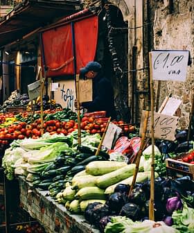 Market stall displaying various vegetables and fruits with price signs in a bustling market setting. - Olive Oil Times