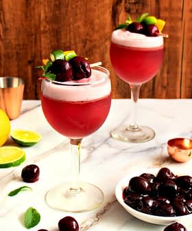 Two cocktails garnished with cherries and lime on a marble surface with citrus fruits and a bowl of cherries. - Olive Oil Times