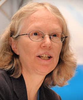 A woman with glasses speaking into a microphone at a conference setting. - Olive Oil Times