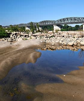 A metal bridge spans a riverbank with sandy terrain and a small pool of water in the foreground. - Olive Oil Times
