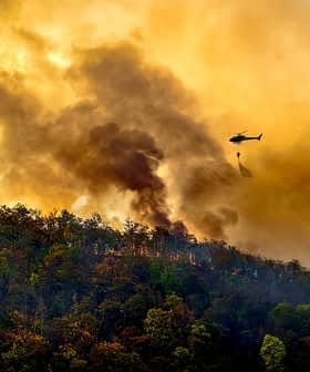 Helicopter dropping water on a forest fire with smoke and flames visible in the background. - Olive Oil Times