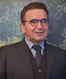 Man wearing a suit and glasses, standing in front of a textured background with greenery. - Olive Oil Times