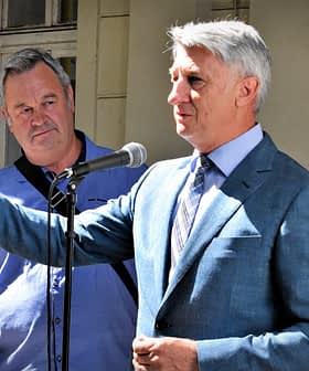 Two men, one speaking into a microphone while the other listens attentively, standing outdoors. - Olive Oil Times