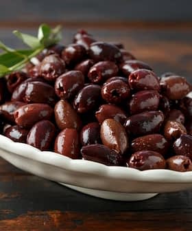 A white bowl filled with a variety of dark olives, garnished with a sprig of olive leaves. - Olive Oil Times