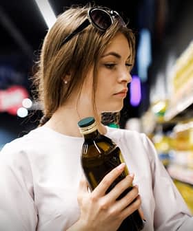 Woman with brown hair holding a bottle of olive oil while examining products on a shelf in a grocery store. - Olive Oil Times