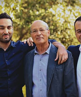 Three men standing outdoors, one with an arm around another, all smiling at the camera. - Olive Oil Times