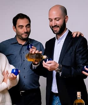 Four individuals holding olive oil bottles and small blue cups in a neutral setting. - Olive Oil Times