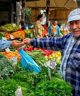 A man in a blue cap interacts with a vendor at a vegetable market filled with various fresh produce. - Olive Oil Times