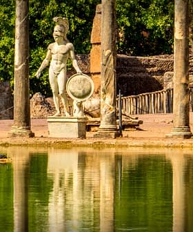 Statue of a Roman warrior holding a shield near ancient columns and water reflection. - Olive Oil Times