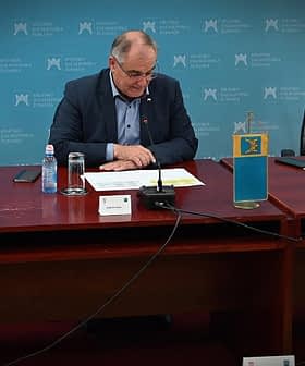 A person sitting at a table with flags of Croatia and another entity displayed on stands. - Olive Oil Times