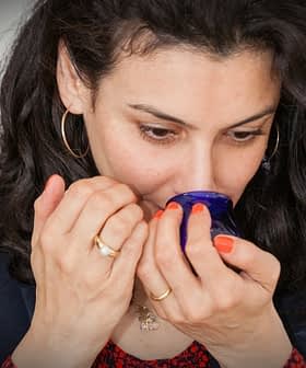 Woman with curly hair smelling from a blue glass container in a neutral setting. - Olive Oil Times