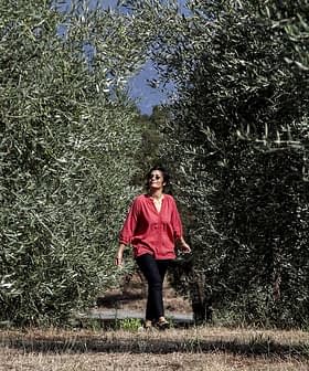 Woman in a red blouse walking between rows of olive trees in an olive grove. - Olive Oil Times