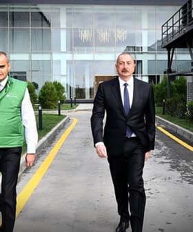 Three men walking on a pathway, two wearing green vests and one in a suit. - Olive Oil Times