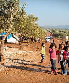 Group of children standing in an olive grove with tents in the background. - Olive Oil Times