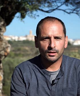 A man sitting in front of a large olive tree, looking directly at the camera with a serious expression. - Olive Oil Times