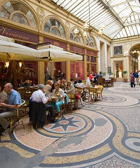 People dining at tables in a covered arcade with a glass roof and decorative floor tiles. - Olive Oil Times