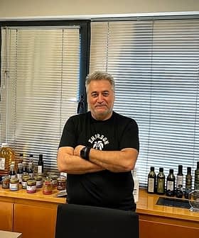 Man with gray hair wearing a black t-shirt standing in an office with olive oil products displayed on a shelf. - Olive Oil Times