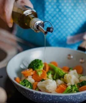 A person in an apron drizzling olive oil over a pan of mixed vegetables including broccoli and carrots. - Olive Oil Times