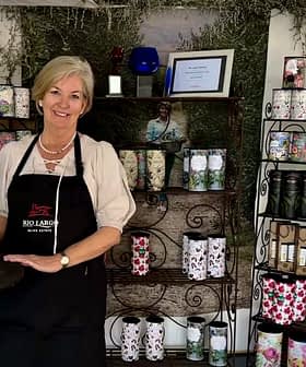A woman wearing an apron stands beside a display of decorative products on shelves. - Olive Oil Times
