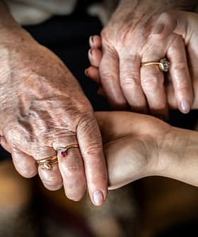 An elderly person's hands holding a younger person's hand, both adorned with rings. - Olive Oil Times