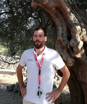 A man wearing a white shirt and lanyard standing beside a large olive tree. - Olive Oil Times